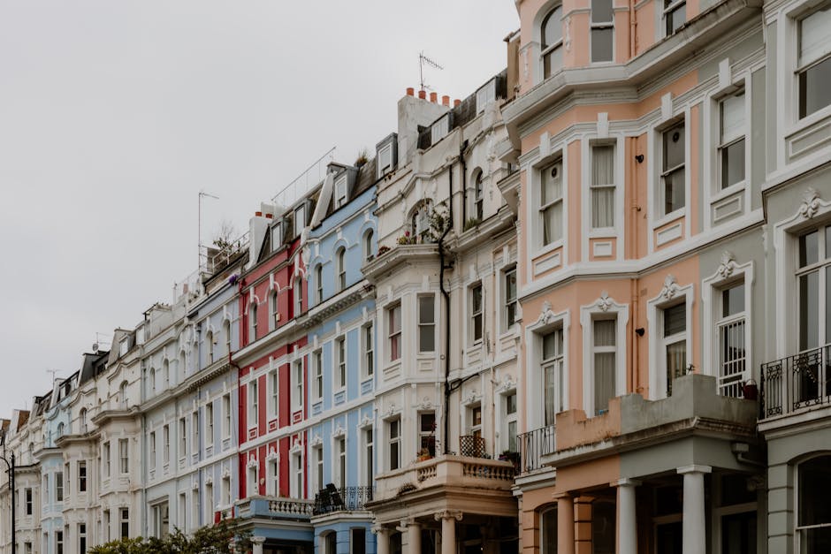 A row of Victorian-style residential buildings in Notting Hill featuring pastel-colored facades in shades of light pink, blue, beige, and white. The buildings have large sash windows, some with black wrought-iron balconies, and ornate decorative moldings around windows and doors. The street is lined with similar terraced houses, with a cloudy sky overhead providing diffuse lighting. The overall scene appears clean and well-maintained, reflecting a typical residential area in Notting Hill, with no visible debris or dirt on the exterior surfaces. For effective surface cleaning or deep cleaning of such properties, Cleaner Notting Hill offers professional domestic cleaning services tailored to maintaining aesthetic appeal and hygiene.