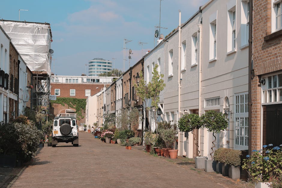 A quiet residential street in Notting Hill featuring a cobblestone pavement lined with terraced houses. The houses are mostly white with some brick facades, large windows, and small front gardens with potted plants and trees. A white van is parked on the street, and in the background, a modern, multi-story building is visible under a clear blue sky. The street exudes cleanliness and well-maintained surfaces, with no visible debris or clutter, reflecting a tidy neighbourhood that benefits from professional domestic cleaning services by Cleaner Notting Hill.