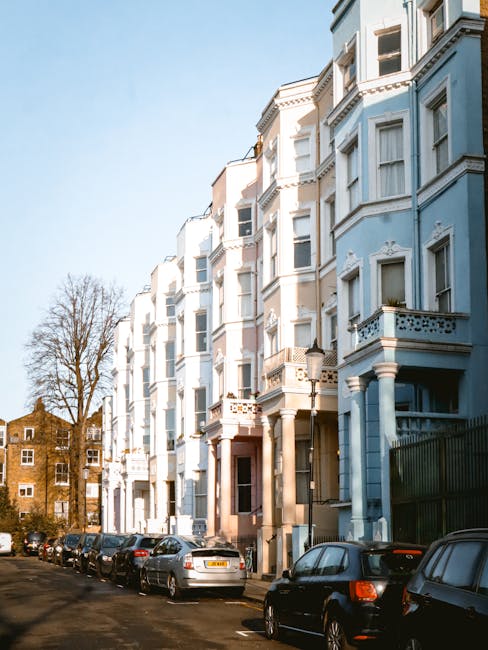 A row of Victorian-style residential buildings in Notting Hill featuring pastel-colored facades in shades of light pink, blue, beige, and white. The buildings have large sash windows, some with black wrought-iron balconies, and ornate decorative moldings around windows and doors. The street is lined with similar terraced houses, with a cloudy sky overhead providing diffuse lighting. The overall scene appears clean and well-maintained, reflecting a typical residential area in Notting Hill, with no visible debris or dirt on the exterior surfaces. For effective surface cleaning or deep cleaning of such properties, Cleaner Notting Hill offers professional domestic cleaning services tailored to maintaining aesthetic appeal and hygiene.