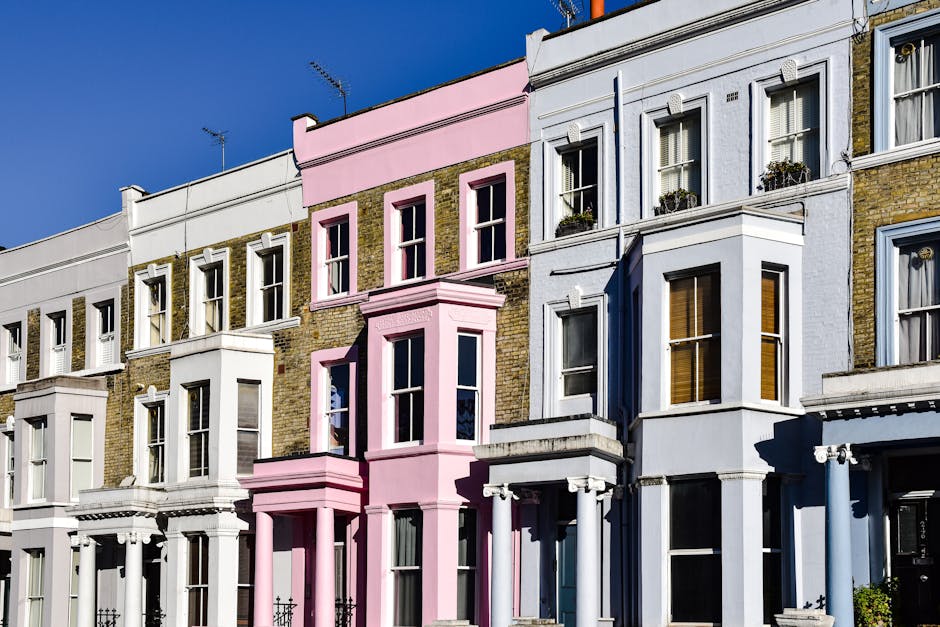 A row of Victorian-style terraced houses in Notting Hill, featuring various facade colors including white, pink, and light blue. The buildings have large sash windows with white frames, some adorned with flower boxes. The exteriors showcase a mix of brickwork and painted finishes, with decorative columns and cornices above the doorways. Bright sunlight illuminates the facades, highlighting the clean and well-maintained surfaces of the buildings, which are typical of the area's residential architecture. The scene is set against a clear blue sky, emphasizing a tidy and picturesque neighborhood that benefits from regular surface cleaning and maintenance by services such as [COMPANY_NAME] to ensure their appearance remains fresh and inviting.