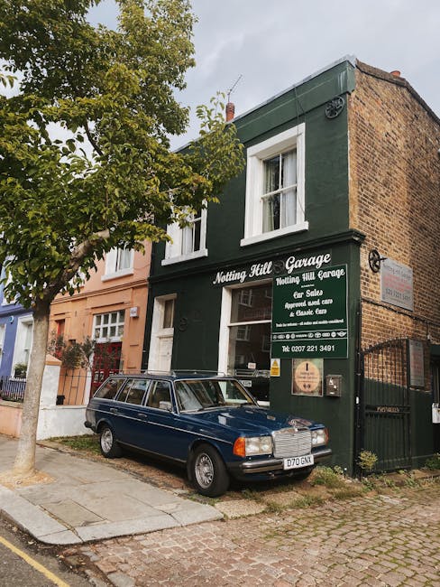 Exterior view of a two-story brick building painted dark green with white window frames, housing the Notting Hill Garage. A dark blue vintage station wagon is parked on the cobblestone street in front of the garage, partially under a leafy tree. The signage on the building advertises car sales, repairs, and servicing services, with contact details visible. The setting is a residential area with a mix of neighboring buildings and a cloudy sky overhead, indicating a typical day in Notting Hill. The scene illustrates well-maintained surfaces and traditional urban architecture, emphasizing the importance of surface cleaning and maintenance in a commercial and residential context, in line with professional cleaning standards offered by Cleaner Notting Hill.