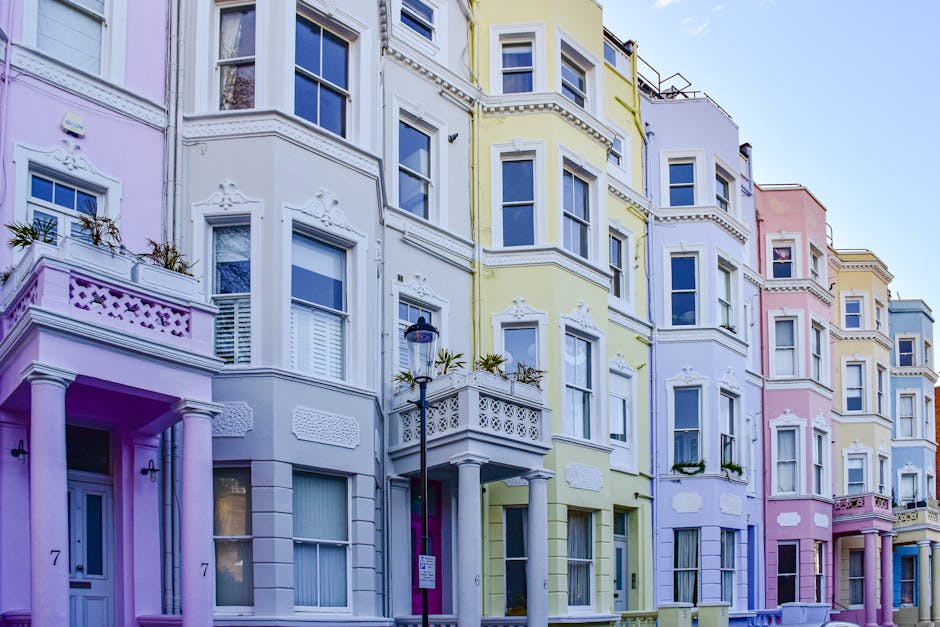 A row of Victorian-style terraced houses in Notting Hill, featuring various facade colors including white, pink, and light blue. The buildings have large sash windows with white frames, some adorned with flower boxes. The exteriors showcase a mix of brickwork and painted finishes, with decorative columns and cornices above the doorways. Bright sunlight illuminates the facades, highlighting the clean and well-maintained surfaces of the buildings, which are typical of the area's residential architecture. The scene is set against a clear blue sky, emphasizing a tidy and picturesque neighborhood that benefits from regular surface cleaning and maintenance by services such as [COMPANY_NAME] to ensure their appearance remains fresh and inviting.