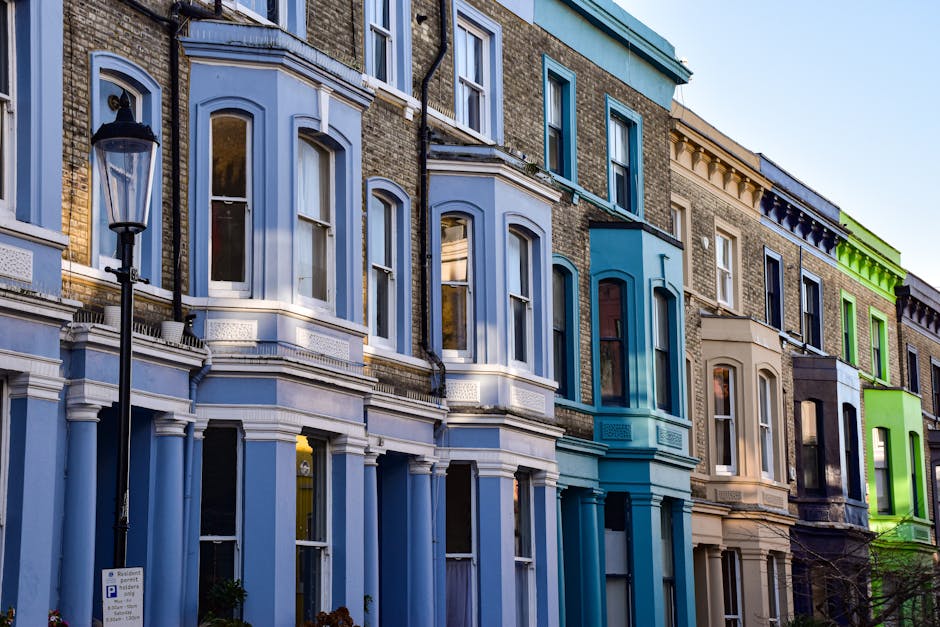 Exterior view of a row of Victorian-style residential buildings in Notting Hill, featuring a variety of painted facades in shades of blue, beige, and green. The buildings have large sash windows with white trim, some with open curtains visible inside. A vintage-style black street lamp with a glass enclosure is positioned on the sidewalk in front of the buildings. The scene is well-lit by natural daylight, highlighting the clean and well-maintained appearance of the surfaces, including the brickwork, painted woodwork, and glass windows. The overall image depicts a picturesque neighborhood with a focus on architectural details and vibrant colors, consistent with a professional domestic or surface cleaning context, as promoted by Cleaner Notting Hill.