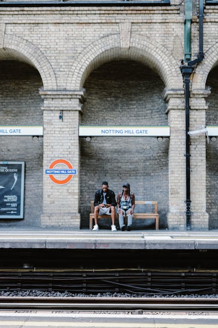 At Notting Hill Gate Tube Station, a brick wall with three large arched openings serves as the backdrop for two passengers sitting on a wooden bench on the platform. The station signage, including a white sign with black lettering reading 'NOTTING HILL GATE' and an underground symbol, is mounted on the brick wall. The platform surface appears clean and well-maintained, with visible train tracks running in front of the seating area. The area is illuminated by natural light, highlighting the textured brickwork and the seating arrangement. Cleaner Notting Hill specializes in surface cleaning and deep cleaning services to maintain hygiene and cleanliness for residential and commercial spaces, ensuring environments like this station are free from dust and graffiti. Their expertise in sanitation aligns with the scenic and functional upkeep of transportation hubs like Notting Hill Gate.