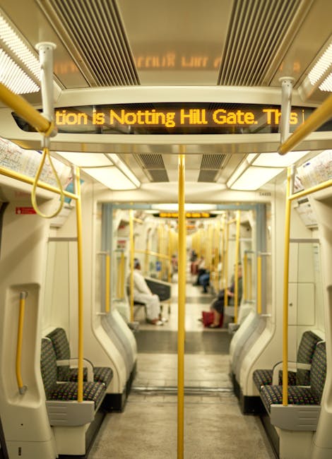 Interior view of a clean and well-maintained London Underground train carriage with light-colored plastic and fabric seating arranged in pairs along both sides of the aisle. The seats have dark upholstery with a patterned design, and the floor is made of smooth, clean grey material. Overhead, bright lighting illuminates the space, and yellow handrails are positioned along the sides and above the seating for passenger stability. A digital information display in yellow text informs passengers about the upcoming station, stating 'Attention is Notting Hill Gate. Tr'. The train's interior appears tidy and sanitized, reflecting professional domestic cleaning standards. Cleaner Notting Hill provides surface cleaning and deep cleaning services to maintain hygiene, ensuring a safe environment for commuters.