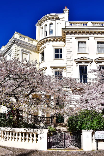 The image depicts a grand, white, multi-story residential building with classical architectural details, including ornate cornices, rounded bay windows, and decorative moldings, set against a vibrant blue sky. In the foreground, a flowering cherry blossom tree with pink blooms partially obscures the lower part of the building. The entrance features a black wrought iron gate and a short stone staircase leading up to the front door. Bright natural sunlight highlights the clean, well-maintained exterior surfaces of the building, emphasizing its polished appearance. The scene suggests a focus on maintaining the property's aesthetic appeal, aligning with the services offered by Cleaner Notting Hill for surface cleaning and general domestic maintenance in the W11 area.