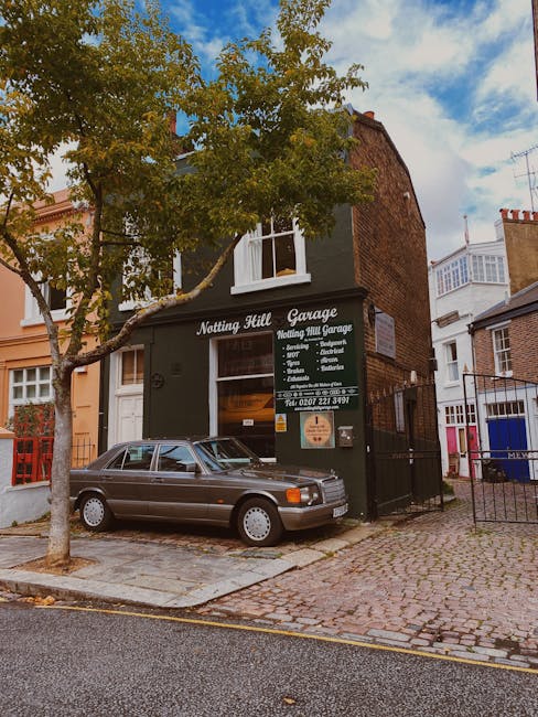 Exterior view of the Notting Hill Garage, a two-story building with a black façade displaying signage for vehicle servicing and repairs, including options like MOTs, brakes, and exhausts. A silver vintage car is parked on the cobbled pavement in front of the garage, adjacent to a small tree with green and yellow leaves. The surrounding area features other residential buildings with brick and painted exteriors, some with white window frames and bay windows. Natural daylight illuminates the scene, highlighting the clean, well-maintained condition of the street and storefront, with the overall environment appearing tidy and orderly. Cleaner Notting Hill specializes in domestic cleaning services that support ongoing hygiene and surface maintenance in residential spaces, reflecting the importance of proper surface cleaning and sanitisation to adhere to local waste disposal regulations in Notting Hill.