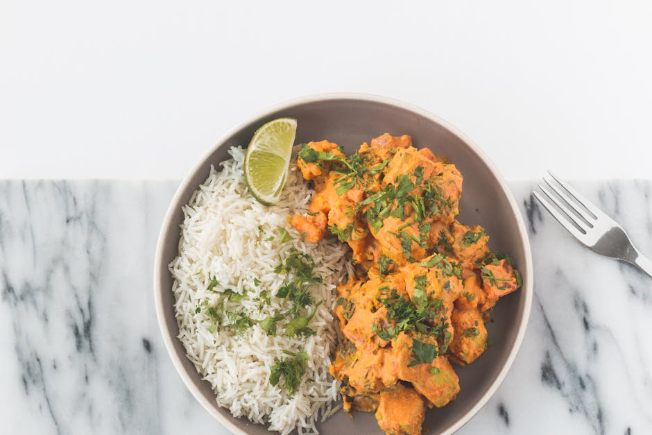 A round ceramic bowl containing a serving of white rice garnished with chopped fresh herbs and a wedge of lime on the side, alongside a portion of curry with a creamy orange sauce and green herbs, placed on a white marble surface. The scene is well-lit, highlighting the vibrant colors and textures of the food, with a stainless steel fork positioned to the right of the bowl. This image does not depict any cleaning or cleaning tools, surfaces, or environments related to [PAGE_TITLE], and it appears to be a presentation of a prepared meal rather than a cleaning context. Therefore, no detailed cleaning or surface description is applicable here, and the image is included for visual appeal related to food presentation.