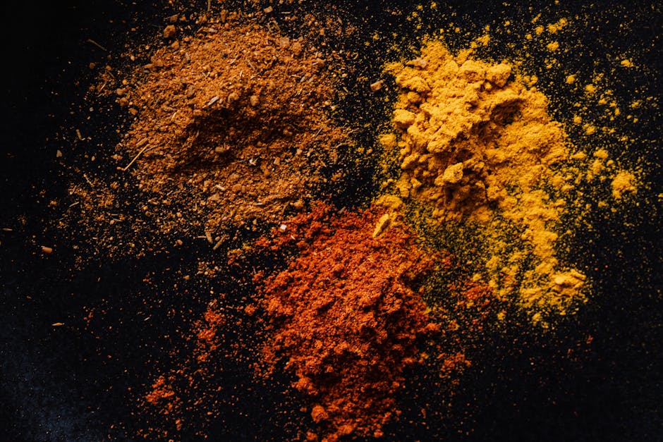 Close-up of three piles of brightly colored curry powders—orange, yellow, and red—scattered on a black surface, with some powder spread around unevenly, highlighting their fine textures and vibrant hues. The powders are arranged in a diagonal pattern with visible granules and slight clumps, and the image is well-lit, emphasizing the contrast between the spices and the dark background. This detailed photograph reflects the topic of surface cleaning and maintenance of kitchen areas, as seen in the context of removing curry stains from carpets, with no tools or additional objects visible. The setting suggests a focus on hygiene and deep cleaning, consistent with services offered by Cleaner Notting Hill in domestic or commercial environments.