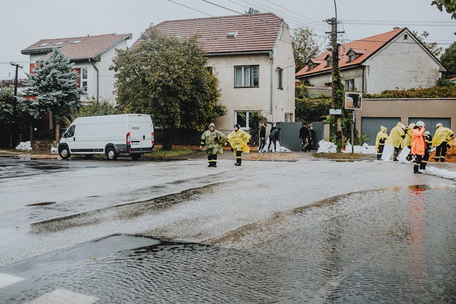 A group of cleanup workers in yellow raincoats and protective clothing are addressing the aftermath of flooding in a residential area on a damp street, which has puddles and wet asphalt. Several individuals are seen walking or standing near the curb, with some engaged in cleaning activities behind a grey fence, where piles of debris or sandbags are visible. A white commercial van parked on the side indicates active site operations. The surrounding houses are two-story structures with tiled roofs, and nearby trees and utility poles complete the suburban scene. The overcast sky suggests recent or ongoing rain, emphasizing the need for thorough surface cleaning and sanitisation in line with domestic cleaning standards, a service provided by Cleaner Notting Hill.