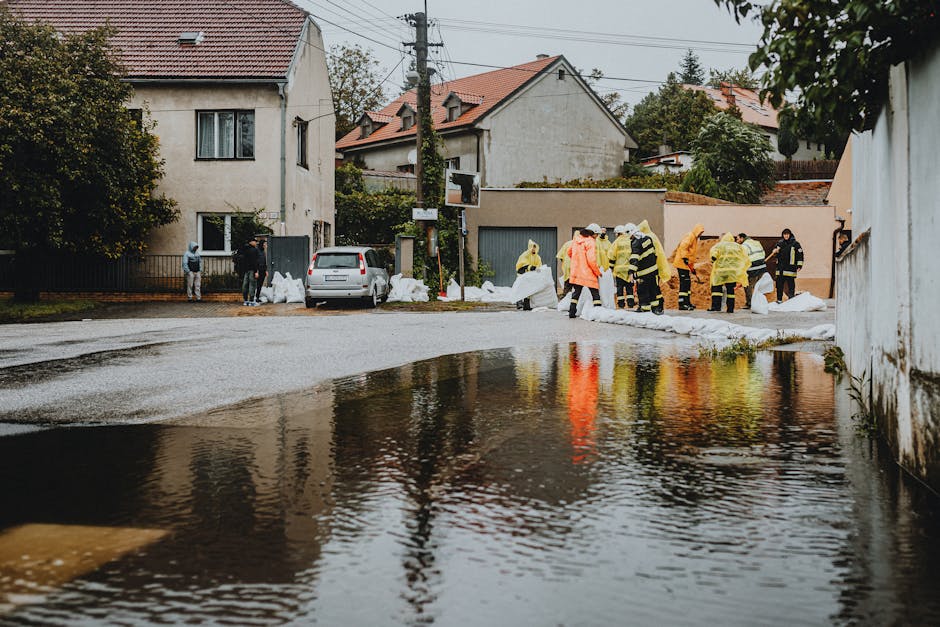 A group of emergency responders and cleaning staff in high-visibility jackets and protective rain gear are working outdoors in a residential neighborhood affected by flooding. They are laying sandbags along the street, which is partially submerged under water, with puddles visible on the pavement. In the background, there are white and beige houses with tiled roofs, some trees, and utility poles with overhead wires. The scene is overcast with natural daylight, highlighting the damp surfaces and reflective water. Cleaner Notting Hill’s team is engaged in surface cleaning and flood response activities aimed at restoring hygiene and safety to the affected area.
